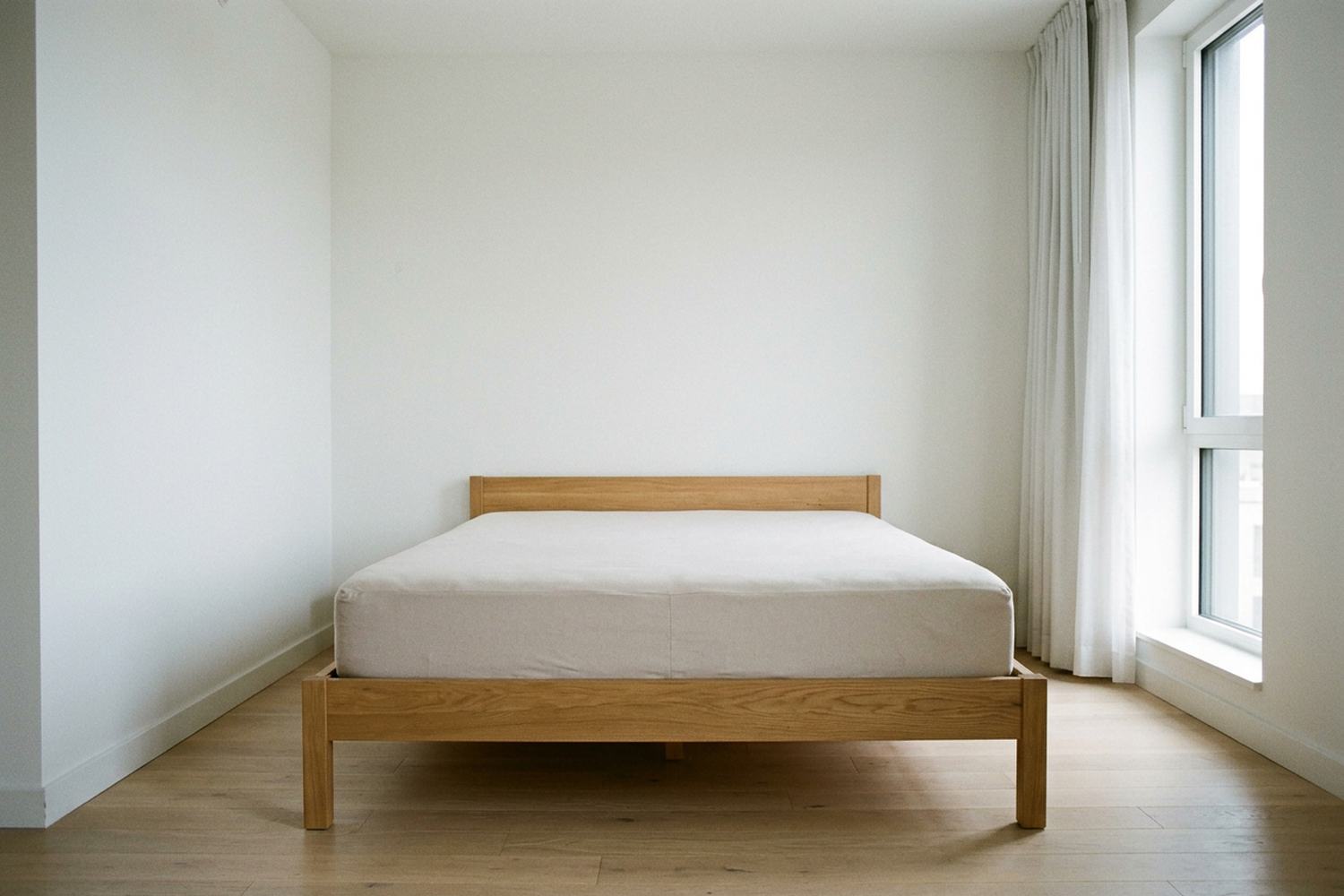 A bare mattress on a pale wood frame in an empty white room beside a tall curtained window.