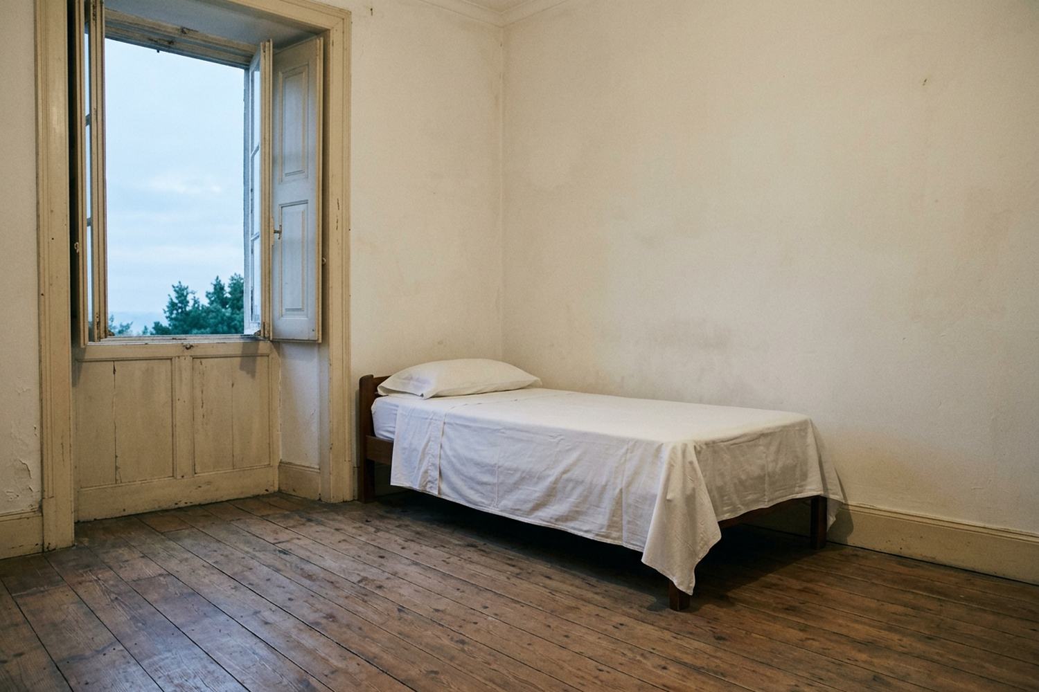 A made bed in a plain room with an open shutter and pale sky beyond.