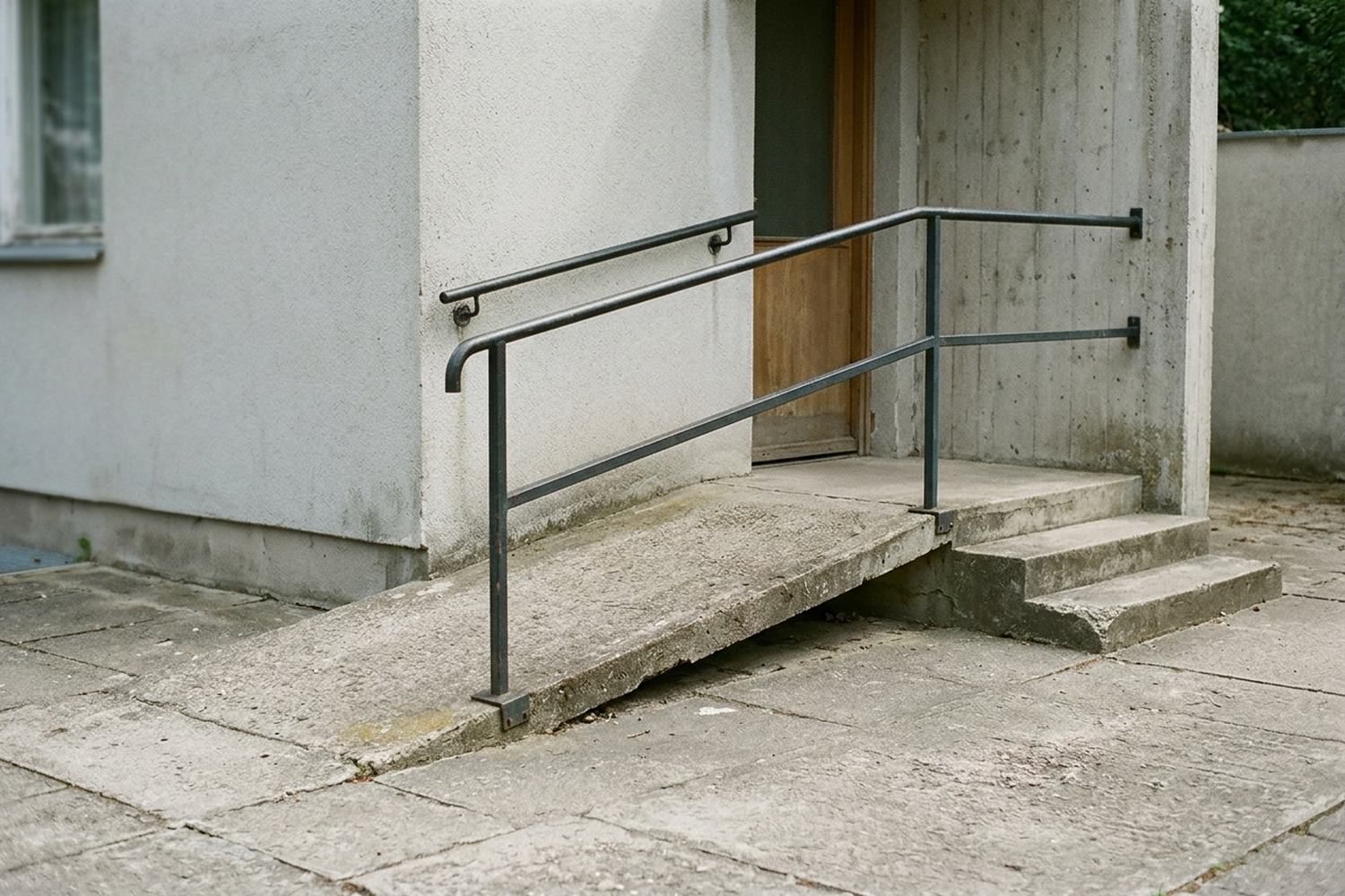 A shallow concrete ramp rising to a threshold beside two low steps, with a rail.